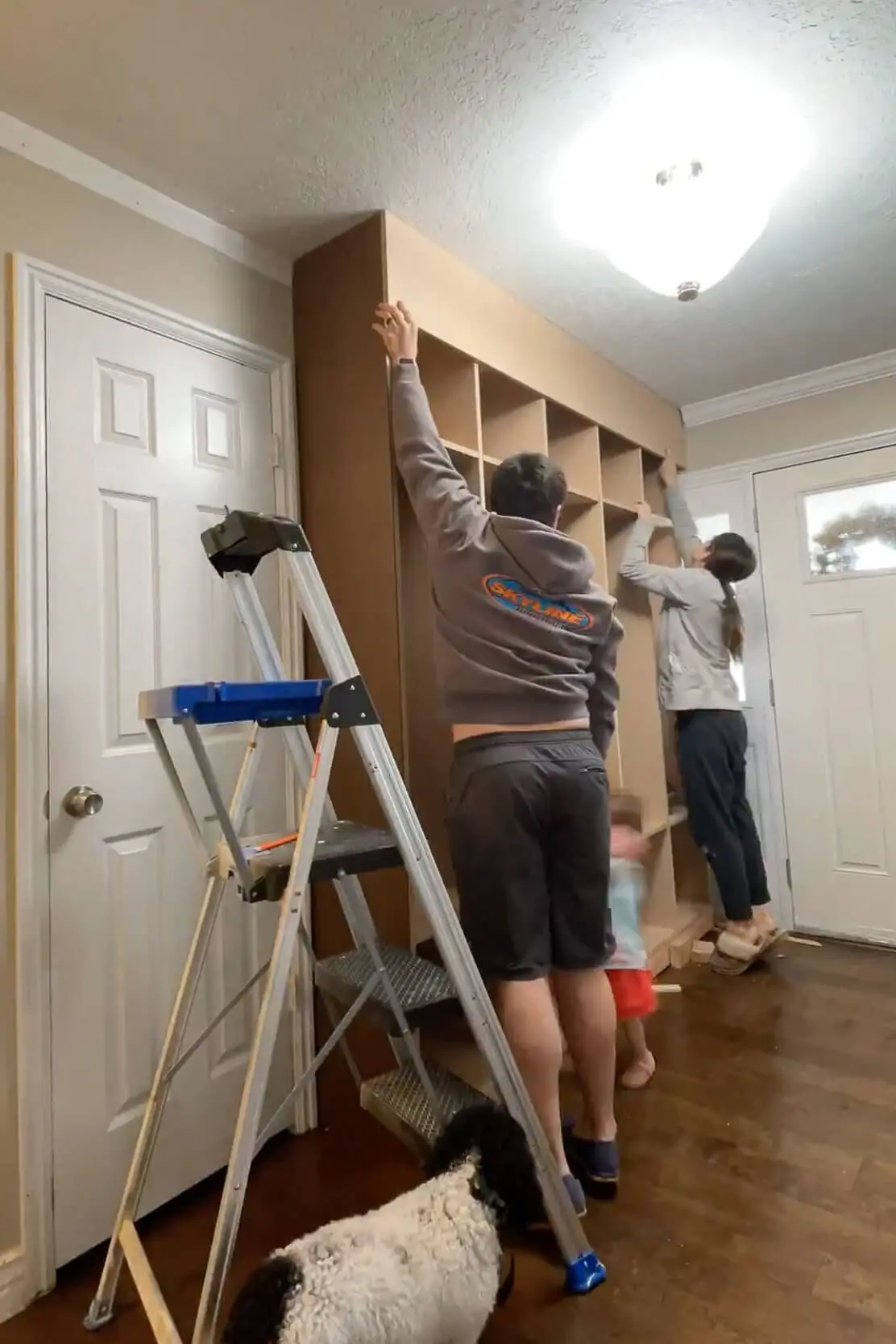 Two people securing a side panel to a custom-built storage unit while a child and dog observe, adding the finishing structure to the project.